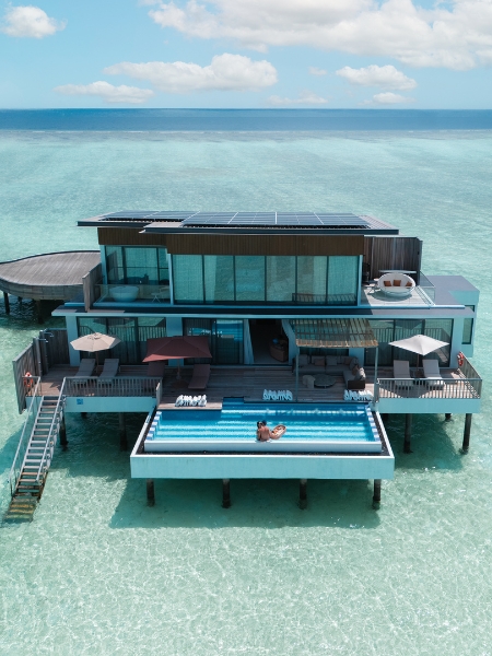 Stunning oceanfront villa with a pool, solar panels, and spacious decks, surrounded by clear turquoise water and a serene sky.