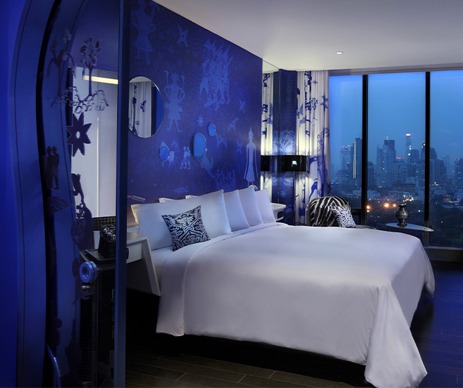 A blue and white themed hotel room with a large bed, desk, and floor to ceiling window displaying a view of Bangkok at night