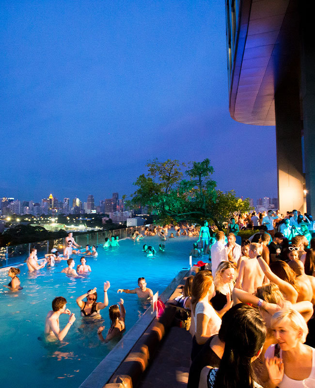 A pool party in an infinity pool overlooking the Bangkok skyline at night. Guests are swimming in the pool and mingling next to it.