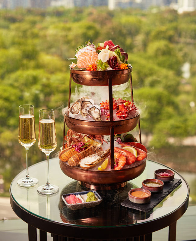 On a table sits a three tiered serving tray filled with seafood, two glasses of wine and condiments. Behind it is a view of Bangkok.
