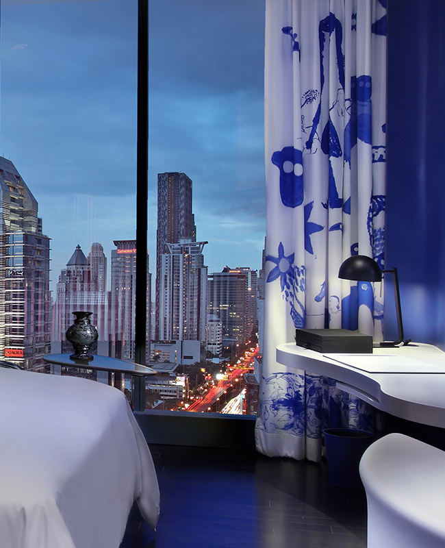 A blue and white theme hotel room with a desk and large floor to ceiling window showing high rise buildings and a busy road below