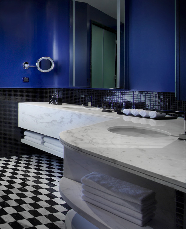 A bathroom with blue painted walls, black marble wall tires and chequered floor. Towels and bath products sit on a large marble sink couter top.