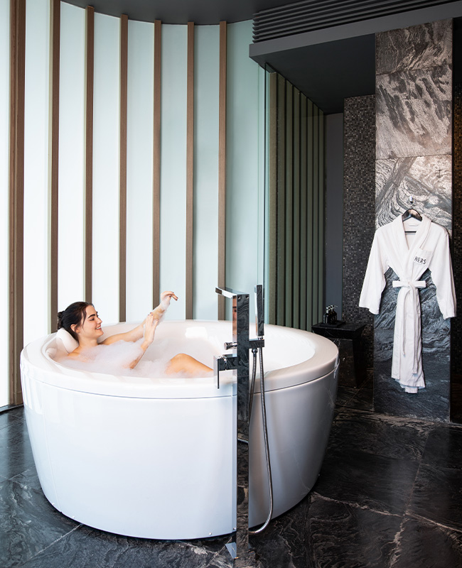 A woman bathes in a round bath tub in a stylish marbled tiled bathroom.