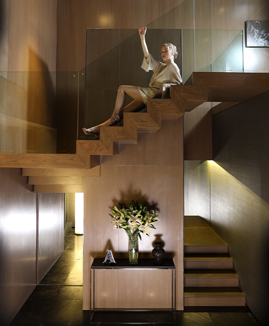 A woman sits posing atop set of open wooden stairs. Below the stairs is a black metal table and vase of flowers.