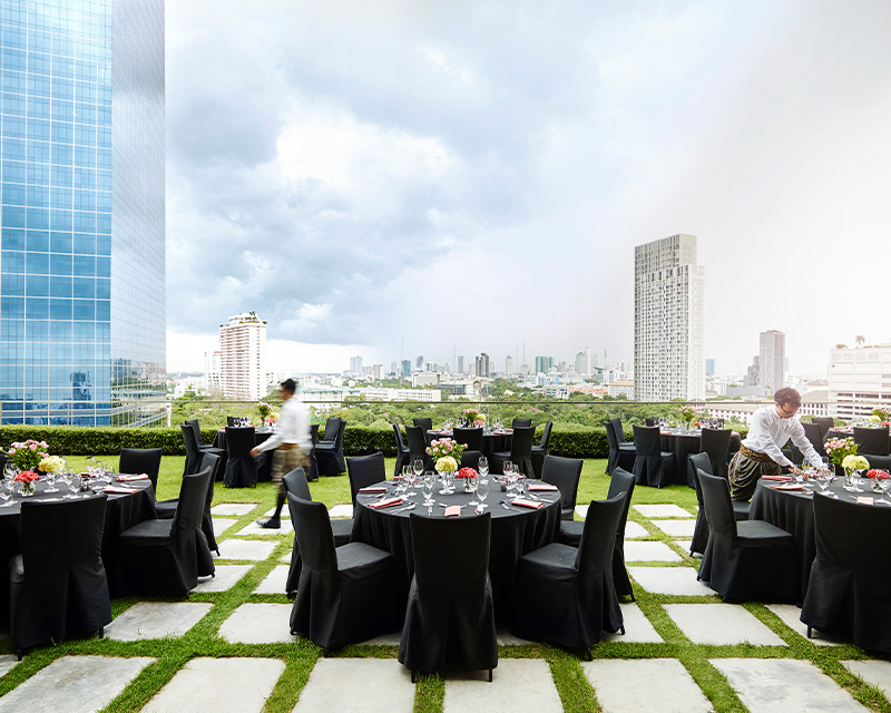 http://Multiple%20round%20dining%20tables%20are%20spread%20out%20on%20a%20grassy%20rooftop%20looking%20over%20the%20Bangkok%20skyline.