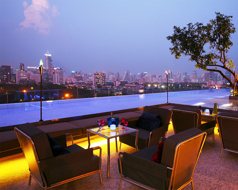 http://A%20large%20orange%20lit%20seating%20area%20stands%20next%20to%20a%20large%20infinity%20pool%20overlooking%20the%20Bangkok%20skyline%20at%20sunset.