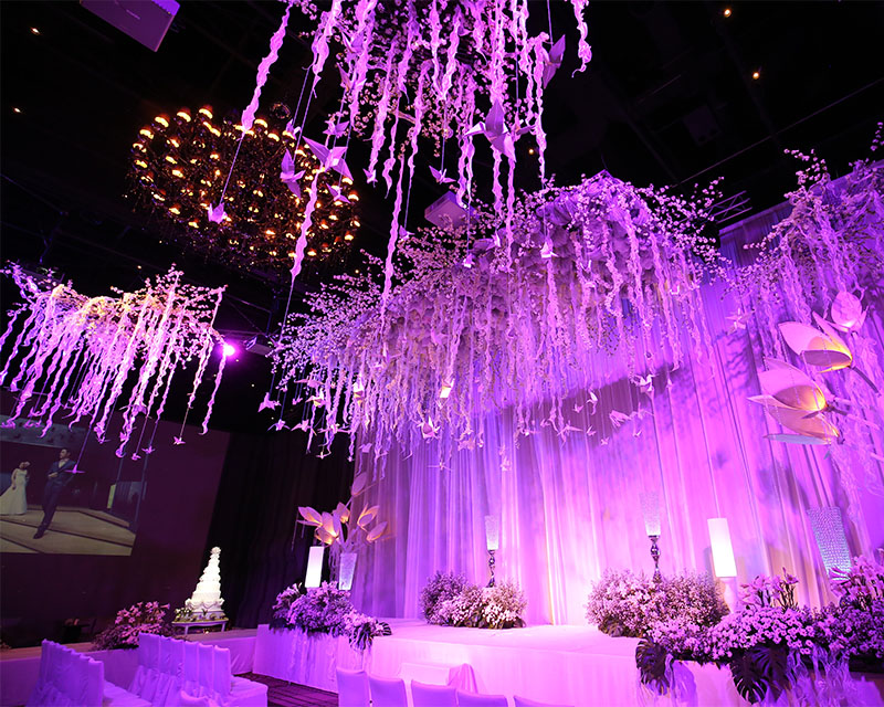 http://A%20large%20ballroom%20with%20ribbons%20hanging%20from%20the%20ceiling%20and%20flowers%20lining%20a%20stage%20are%20lit%20up%20with%20a%20purple%20glow.