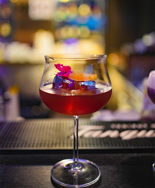 A delicious looking red cocktail sits on a bar.