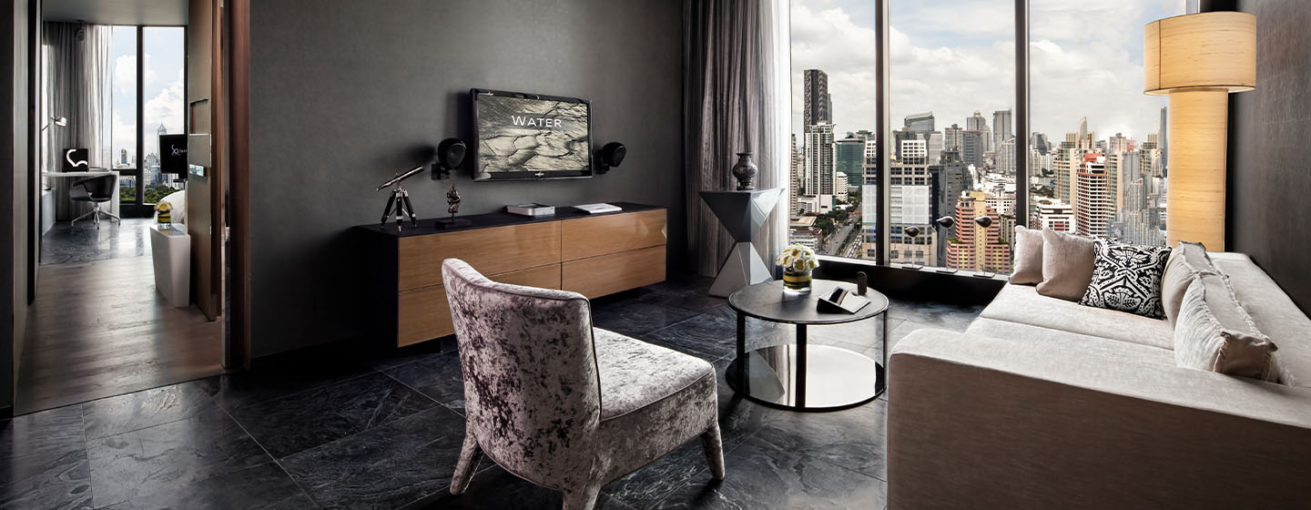 A hotel room living space with a large couch, cushioned chair, small coffee table and mounted flatscreen TV. Floor to ceiling windows display a beautiful skyline view.