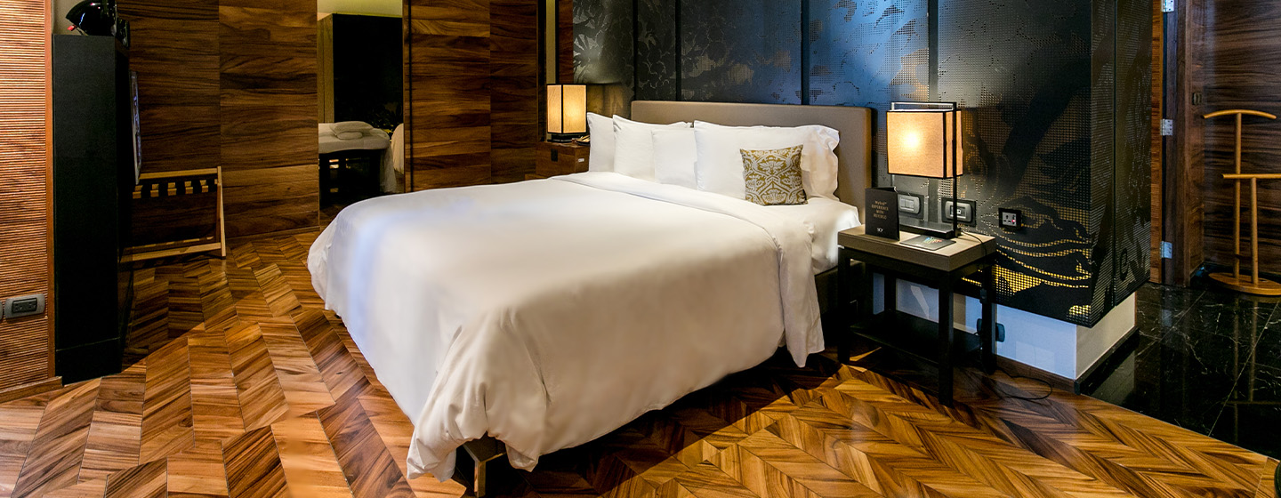 A large hotel bedroom with wooden floors and walls. A large bed sits infront of a black feature wall, either side are bedside tables with lamps.