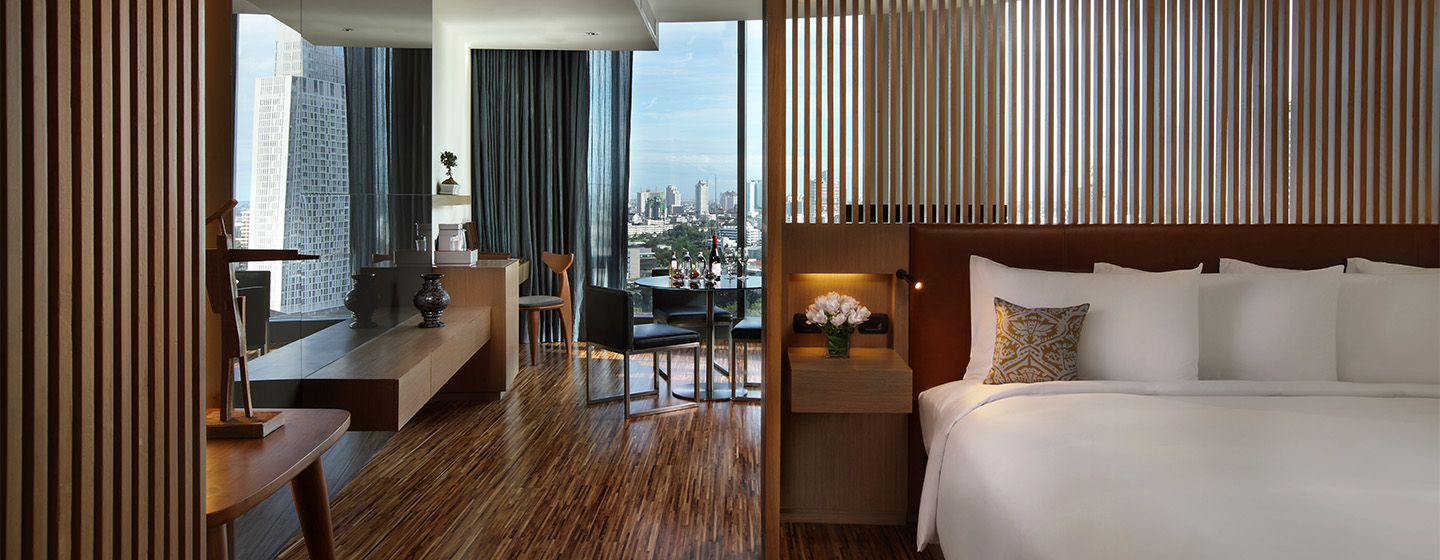 An open, wood themed hotel room. A wooden slatted wall separates a large bed and a small dining area. Floor to ceiling windows overlook Bangkok.