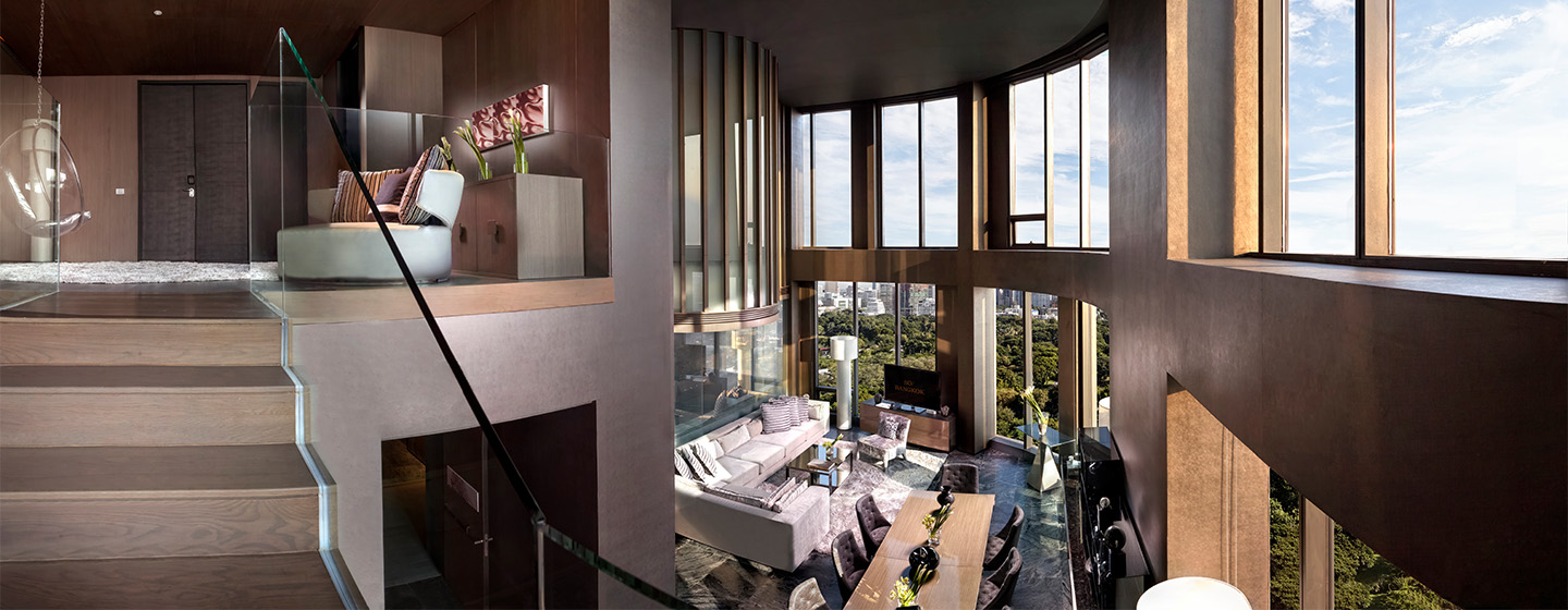An elevated view of a large, multi level hotel suite surrounded by windows. On the ground floor is a large corner couch, TV stand and large dining table. A staircase leads up to another small seating area.