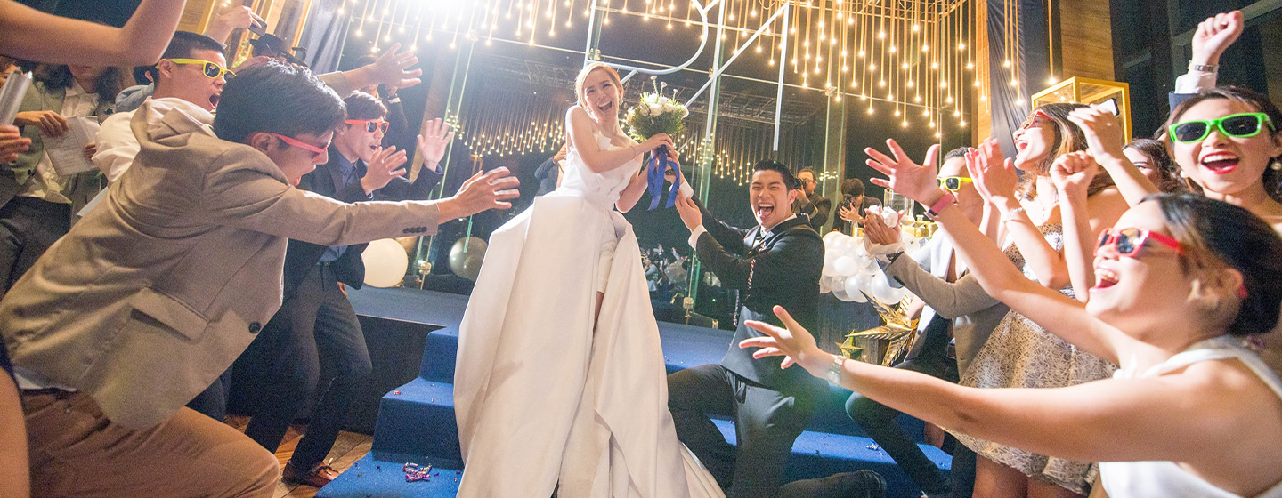 A woman in a wedding dress and a man in a suit are being celebrated by friends underneath beautiful gold lights.