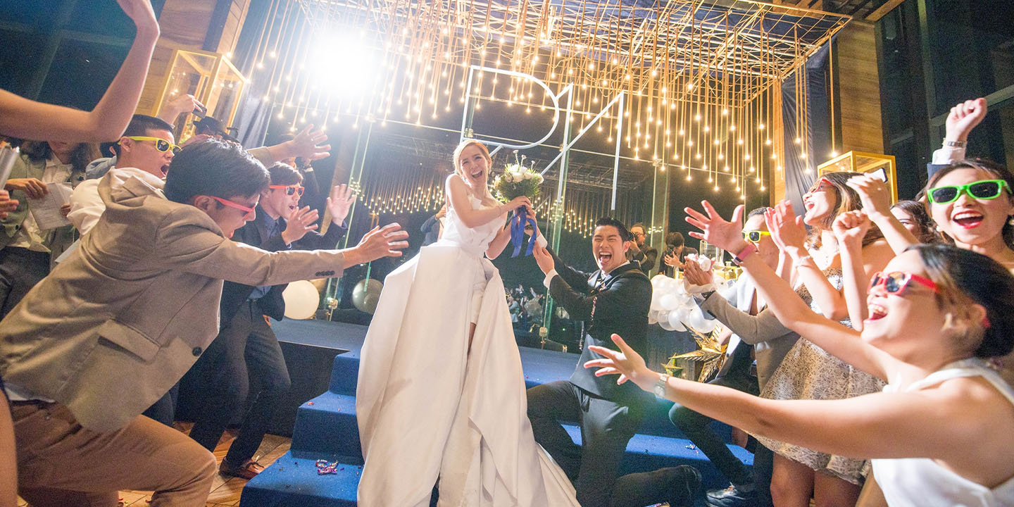 A woman in a wedding dress and a man in a suit are being celebrated by friends underneath beautiful gold lights.