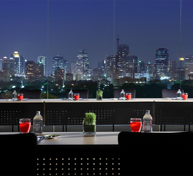 Tiered meeting tabled with glasses, water bottles and plants sit by a floor to ceiling window overlooking the Bangkok skyline at night.