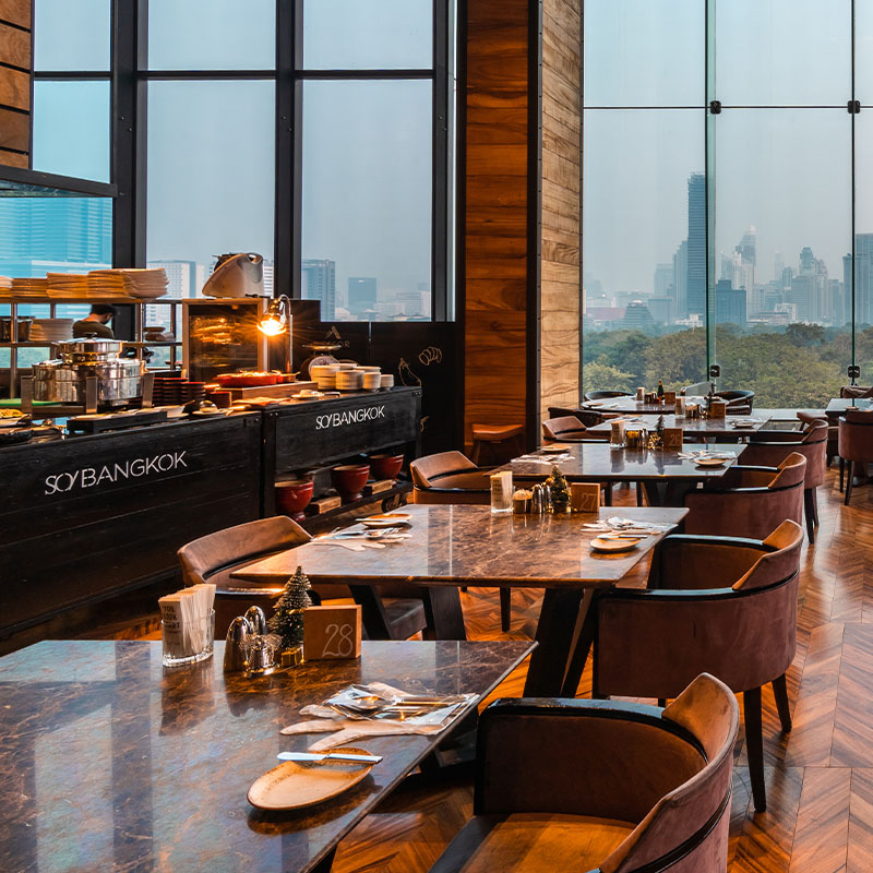 A large restaurant. There are multiple dining tables and chairs and a buffet table and floor-to-ceiling windows overlook Bangkok's Lumpini Park.