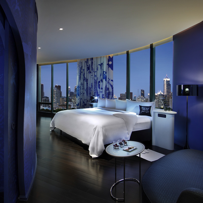 A large, blue hotel room. A king bed sits by floor to ceiling windows overlooking Bangkok at night. An archway leads from the room to a coffee station.