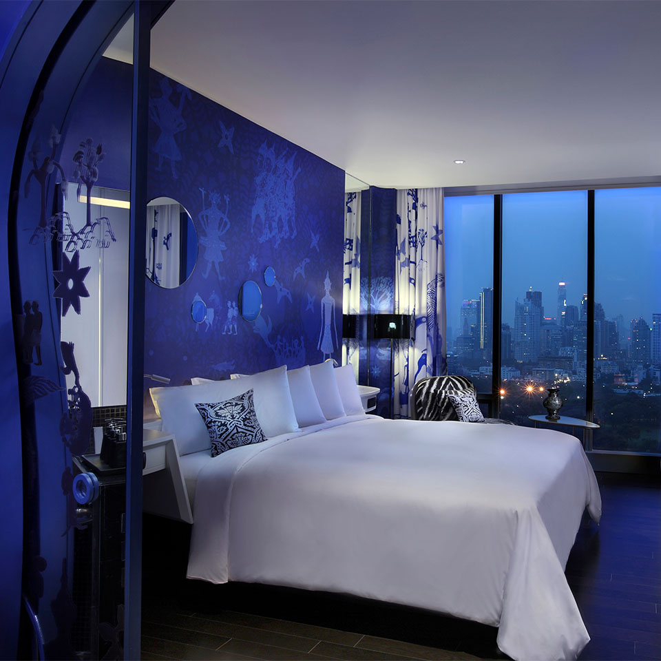 A blue and white themed hotel room with a large bed, desk, and floor to ceiling window displaying a view of Bangkok.