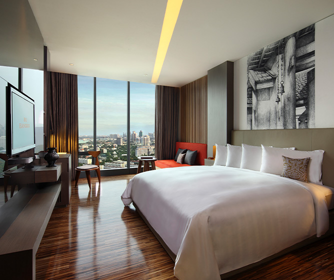 A wooden themed hotel room. A king bed sits in the middle opposite a large TV and a seating area overlooks a view of Bangkok.