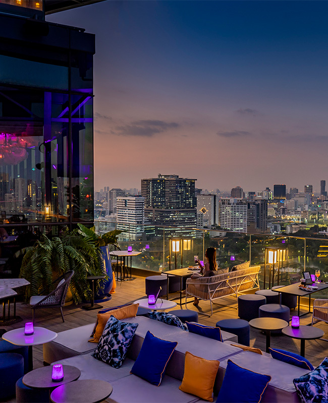 A large rooftop seating area bathed in a purple glow overlooks the Bangkok slyline at sunset
