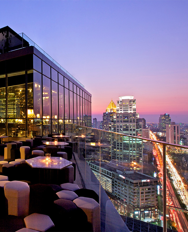 A large, candle lit rooftop bar seating area overlooks a view of a busy Bangkok at sunset