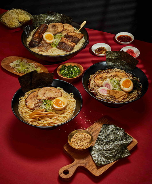 http://Three%20large%20bowls%20of%20ramen%20with%20a%20variety%20of%20condiments%20and%20toppings%20in%20small%20bowls%20around%20the%20table.