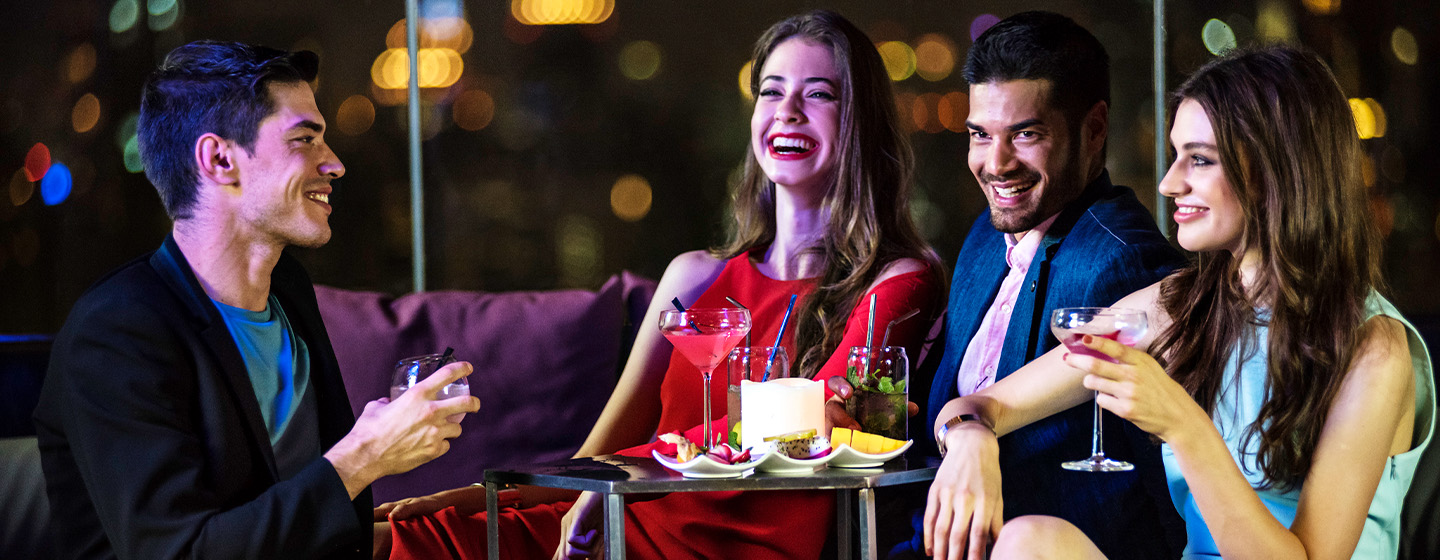 Four people are sitting in a rooftop bar area at night enjoying cocktails food.