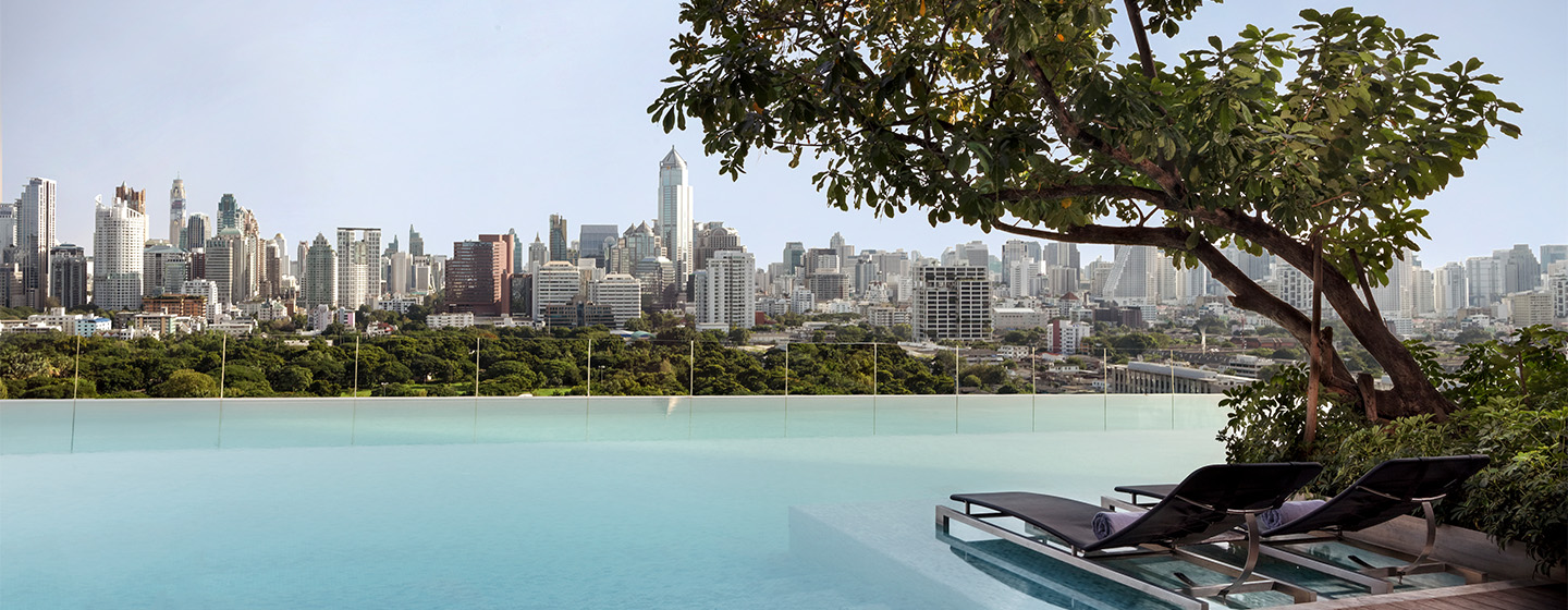 Two sunbeds sit, partially submerged in an infinity pool, underneath a large overhanging tree. The pool overlooks the Bangkok skyline.