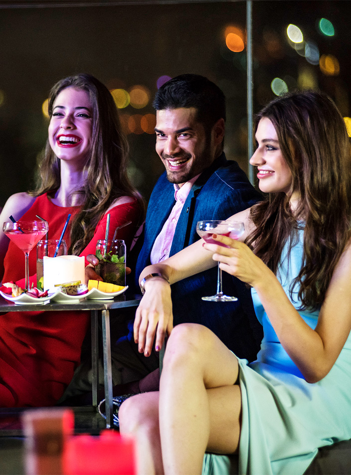 Four people are sitting in a rooftop bar area at night enjoying cocktails food.