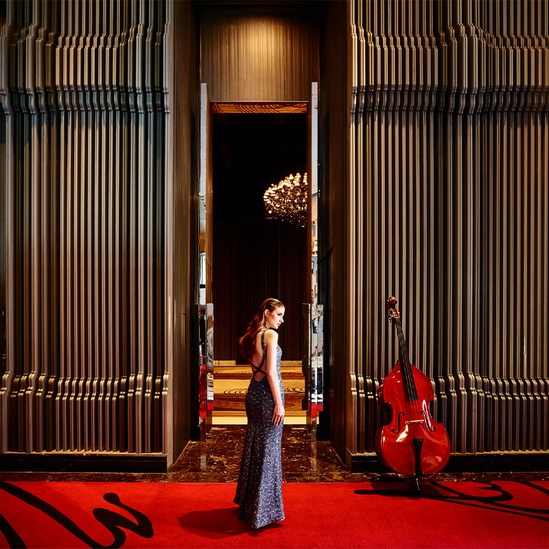 A woman in a blue dress stands in front of a door way. A cello leans on a bronze textured wall.