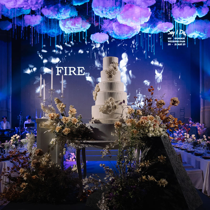A large room with dining tables bathed in blue and purple light A tiered wedding cake stands on a separate table surrounded by flowers.