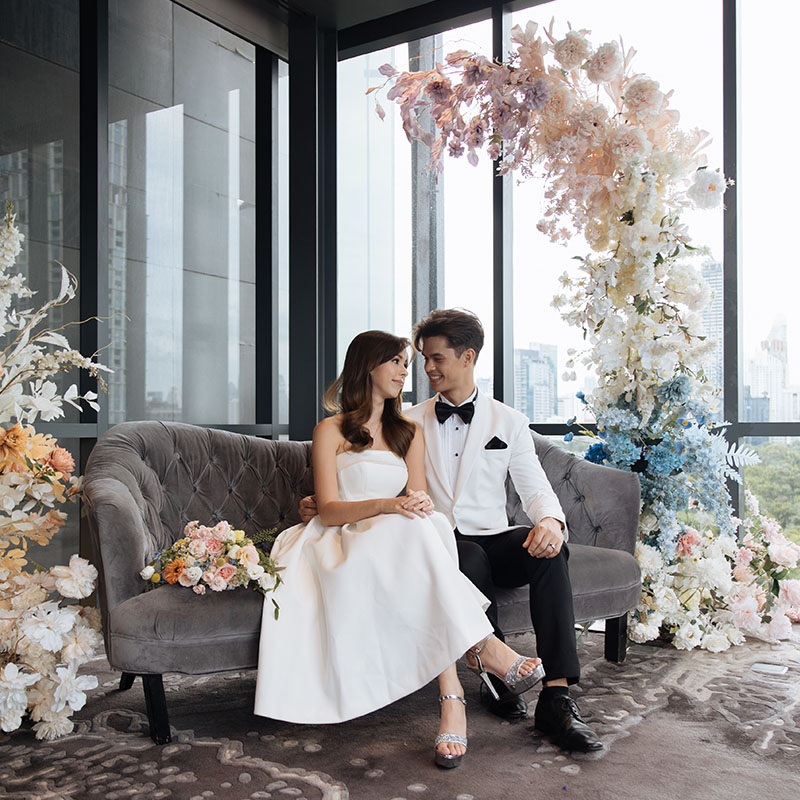 A man and women dressed in wedding outfits sit on a grey couch. A beautiful flower arrangement towers above them in front of a floor-to-ceiling window.