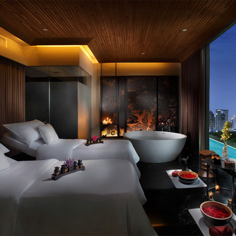 Two spa loungers and a bathtub sit in a large room. Floor to ceiling windows look out onto a outdoor pool, park and skyline.