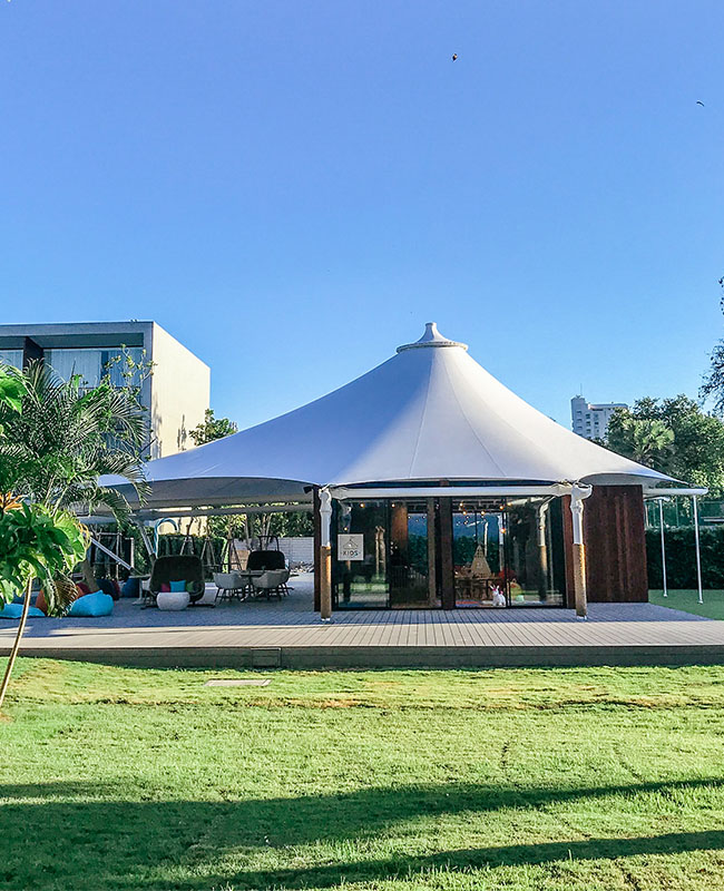A large white tent with wooden pillars stands on a lush green lawn. The setting is bright and sunny, surrounded by trees and a modern building.