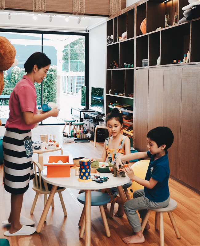 A woman engages with two children at a small round table in a playroom. The atmosphere is warm, with toys scattered and a shelf filled with items.
