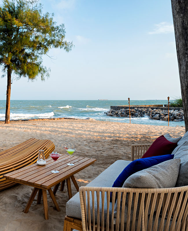Cozy beach scene with a wooden lounge set under a tree, overlooking gentle waves and a distant pier. Relaxing, serene atmosphere with soft sand.
