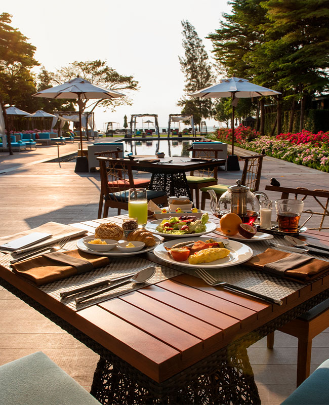 Outdoor dining scene with a wooden table set for breakfast, featuring pastries, fruit, and juice. Sunlit path leads to a scenic, poolside view. Relaxed atmosphere.