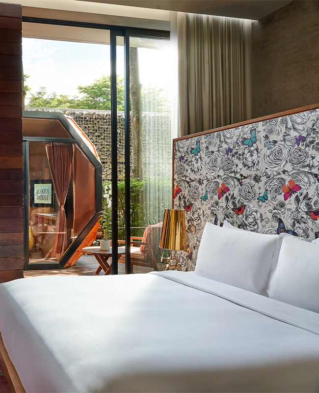 Modern hotel room with a large bed, patterned butterfly wallpaper, and a cozy seating area. Glass doors lead to a lush outdoor patio, creating a serene ambiance.