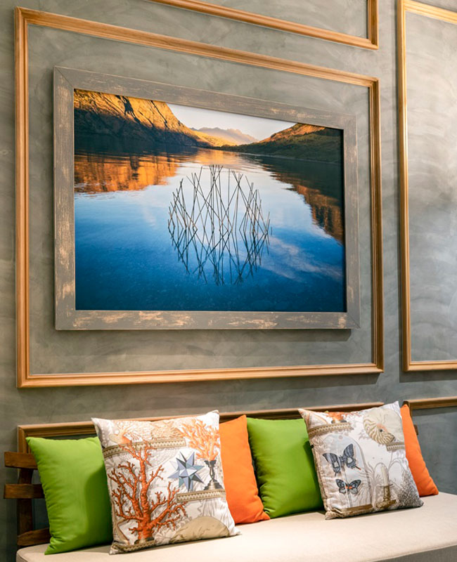 A framed painting of a serene lake with mirrored mountains hangs on a wall. Below are colorful pillows with nature motifs on a bench.