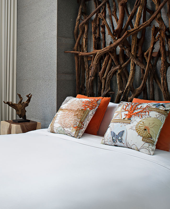 Modern bedroom with a white bed, colorful pillows, and a unique headboard made of intertwined branches. Sunlight streams through large glass doors.