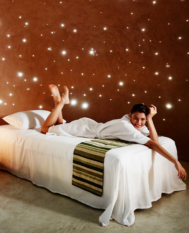 A person in a white robe lounges on a bed with white linens against a warm brown wall adorned with twinkling lights, creating a serene and cozy ambiance.