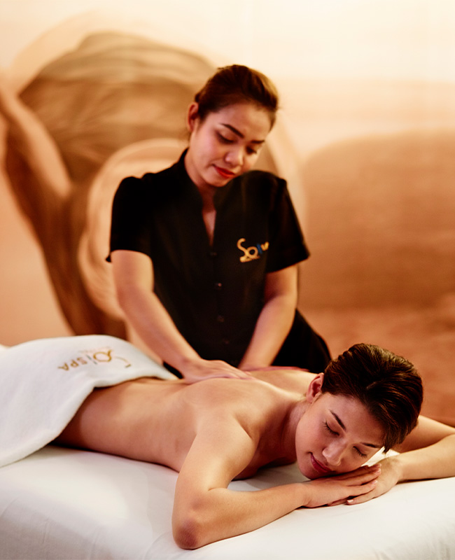 A woman in uniform gives a relaxing back massage to a person lying face down on a massage table in a serene spa setting, creating a soothing atmosphere.