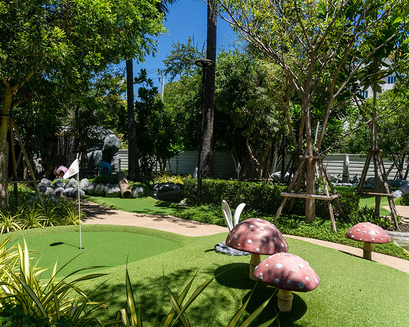 http://A%20sunny%20mini-golf%20course%20with%20large,%20whimsical%20red%20mushrooms%20on%20artificial%20grass.%20The%20course%20is%20bordered%20by%20lush%20trees,%20creating%20a%20playful%20atmosphere.