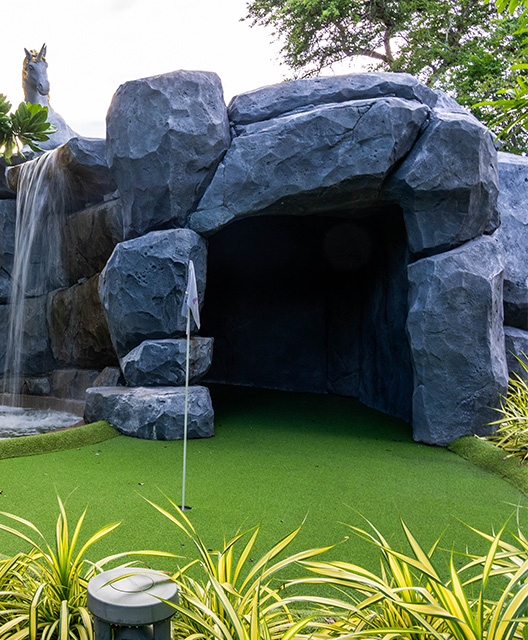 http://A%20mini-golf%20course%20entrance%20featuring%20a%20cave%20with%20large,%20textured%20rocks.%20Green%20artificial%20turf%20leads%20inside,%20surrounded%20by%20lush%20plants,%20creating%20a%20playful,%20adventurous%20tone.