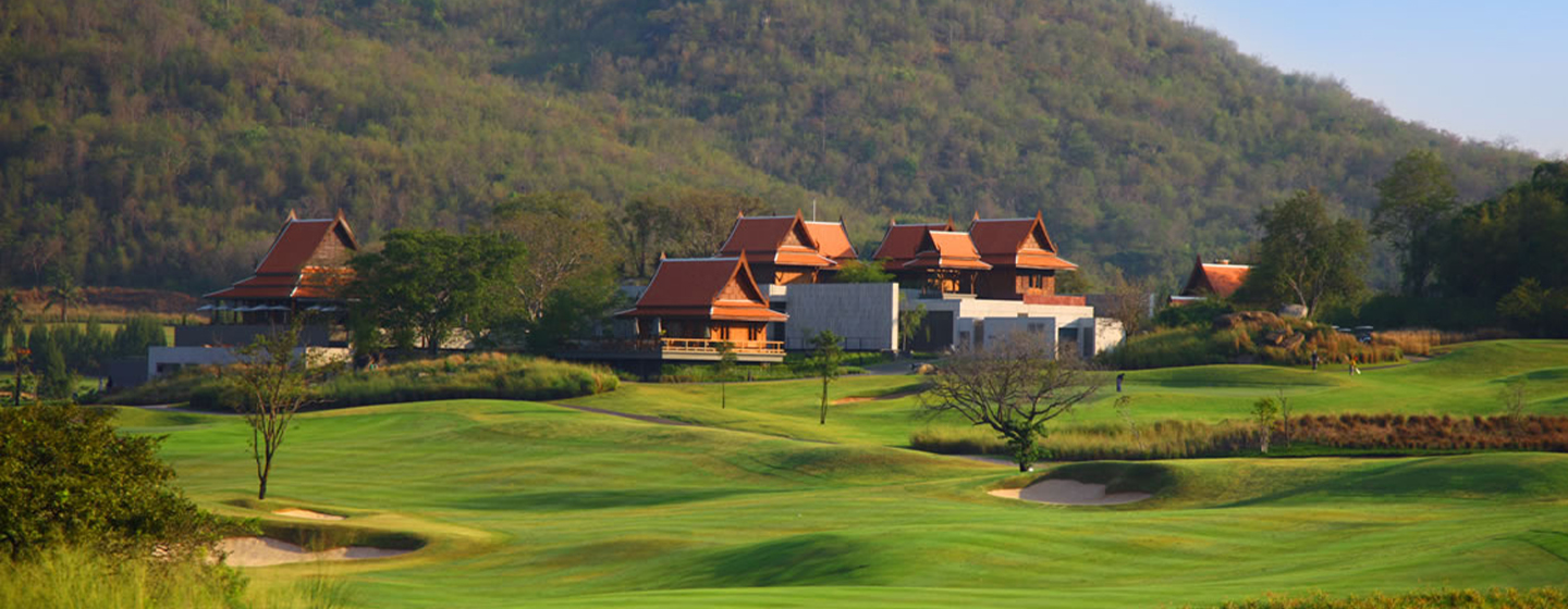 Scenic view of a lush green golf course with rolling hills, traditional-style buildings with red roofs, and wooded hills in the background under a blue sky.