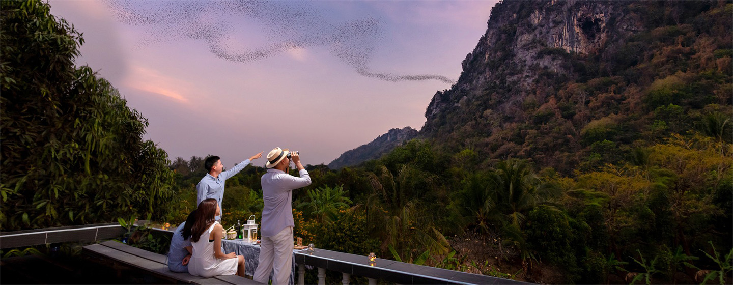 Three people watch a flock of bats emerging from a cave in a mountain at dusk. The sky is purple, with lush greenery surrounding the scene, creating a serene atmosphere.