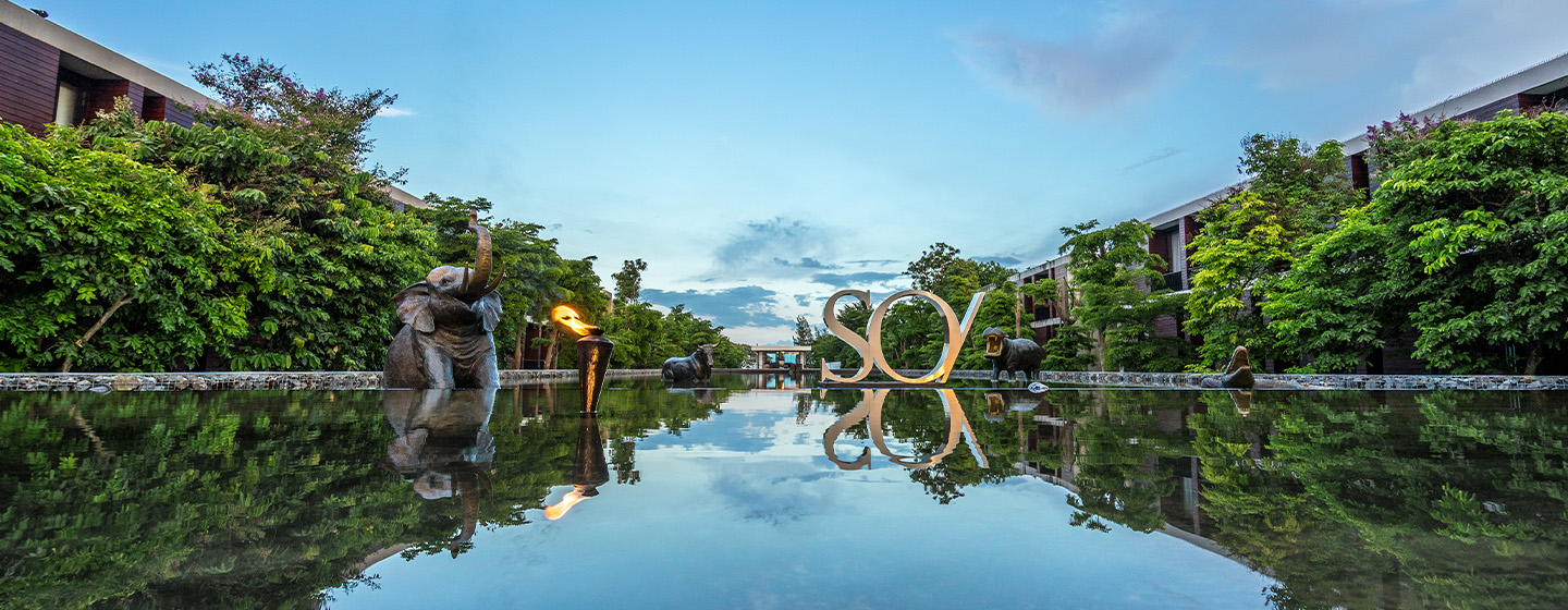A serene water feature reflecting a clear blue sky, flanked by lush greenery. Elephant sculptures and a lit torch add elegance to the tranquil scene.