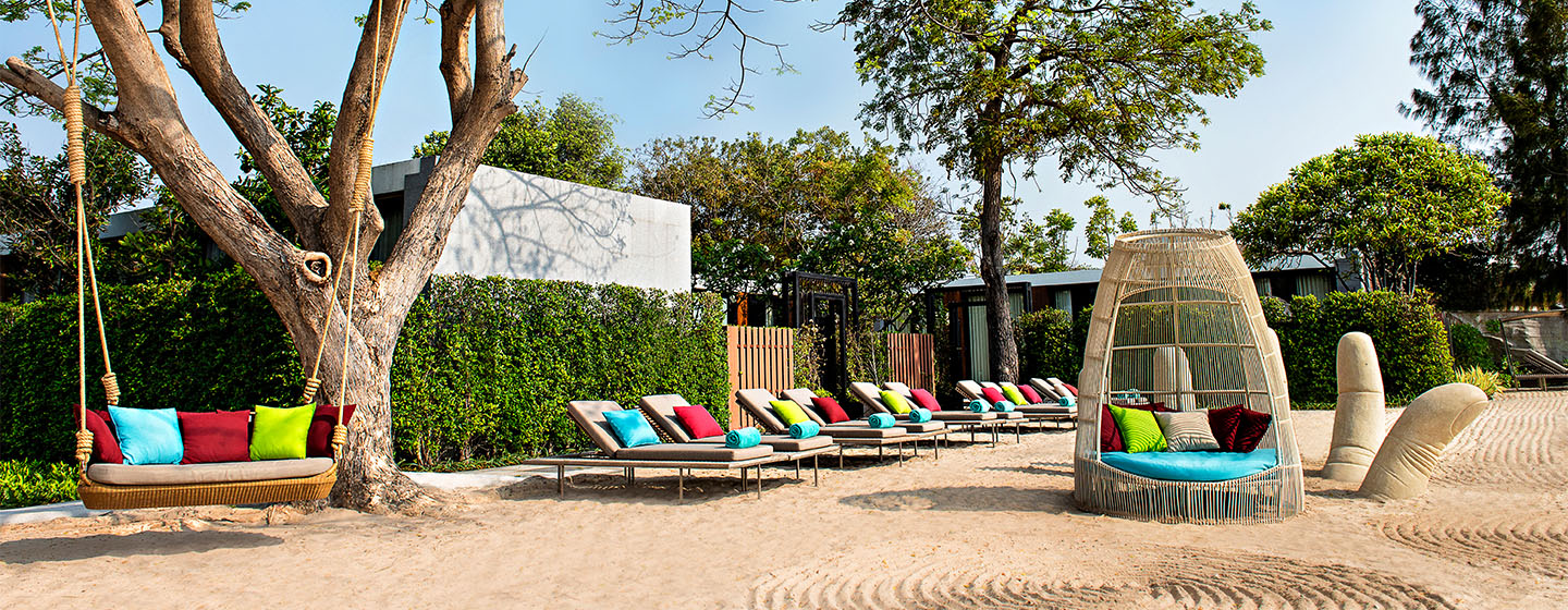 A serene outdoor lounge area with colorful cushioned chairs under a tree. Nearby, a woven seating pod and greenery create a relaxed, inviting vibe.