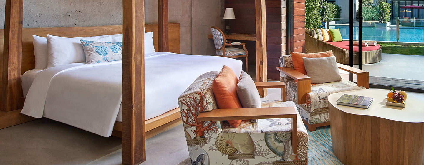 Luxurious bedroom with a large bed, wooden furniture, and cozy armchairs. A glass door opens to a vibrant pool area, creating a serene and inviting atmosphere.