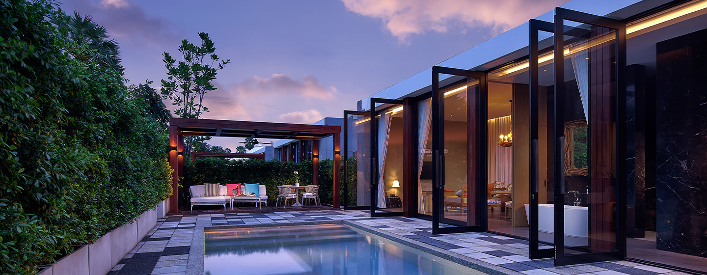 Modern house exterior at dusk with open glass doors, a lit interior, pool in the foreground, and greenery surrounding the patio area, creating a serene atmosphere.